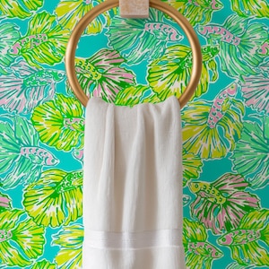 May include: A white towel hangs on a gold ring towel rack against a turquoise background with a repeating pattern of green and pink fish.