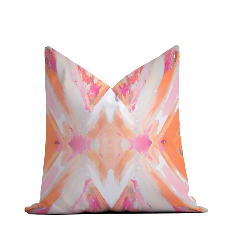 May include: A decorative pillow with a geometric pattern in shades of pink, orange, and white. The pillow has a white background with a diamond-shaped pattern in the center. The diamond is surrounded by a border of pink and orange.
