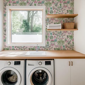 May include: A laundry room with a light pink wallpaper featuring a tropical design. The room includes a white sink, two wooden shelves, a window, and two white washing machines. A wooden countertop runs along the wall.