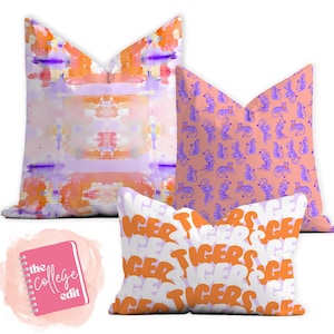 May include: Three decorative throw pillows with a college theme. The first pillow has a watercolor design in shades of orange, pink, and purple. The second pillow has a repeating pattern of purple tigers on a peach background. The third pillow is white with the word "TIGERS" repeated in orange.