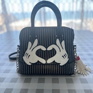 May include: Black and white striped handbag with a white heart shape made from Mickey Mouse hands. The bag has a white tassel.