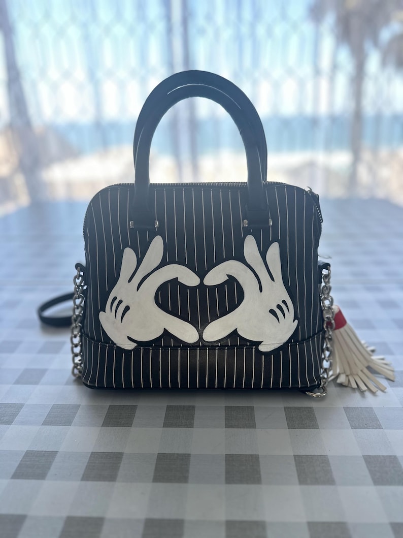 May include: Black and white striped handbag with a white heart shape made from Mickey Mouse hands. The bag has a white tassel.