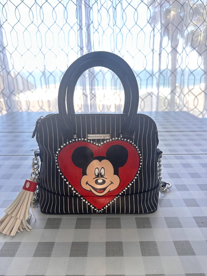May include: Black and white striped handbag with a red heart featuring a cartoon image of Mickey Mouse. The bag has a black handle and a chain strap. A white tassel hangs from the zipper pull.
