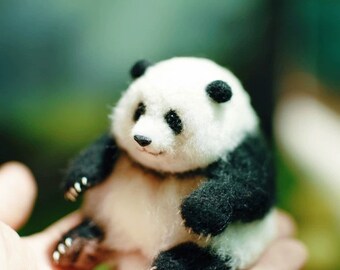 May include: A small, white and black panda bear plush toy with black eyes and a black nose. The panda is sitting on a hand.