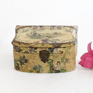 May include: An oval-shaped decorative box with a hinged lid. The box is beige with a floral pattern of blue and white pansies. The lid features a pastoral scene. A decorative metal clasp secures the box.