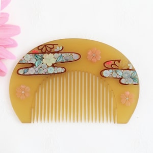May include: A yellow comb with a curved top and fine teeth. The comb is decorated with floral designs in shades of pink, blue, and white, set against a brown outline. The comb has a traditional Japanese aesthetic.