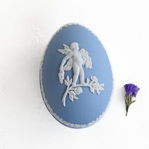 May include: A blue oval-shaped decorative box with a white cherub and floral relief design. The box has a white trim and is next to a small purple flower on a white background.