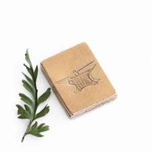 May include: A small, rectangular, gold-colored metal box with an embossed anvil design on the top. A sprig of green foliage is positioned near the box against a white background. The box appears to be a small container or case.