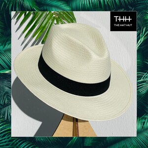 Pu&ograve; includere: Un cappello di paglia bianco con una fascia nera. Il cappello &egrave; appoggiato su un supporto in legno. Il testo "THH THE HAT HUT" &egrave; nell'angolo in alto a destra dell'immagine.