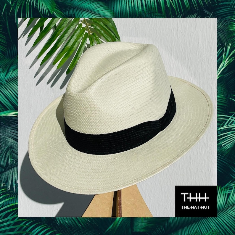 Pu&ograve; includere: Un cappello Panama bianco con una fascia nera. Il cappello &egrave; fatto di paglia intrecciata e ha un'ampia tesa. Il cappello &egrave; appoggiato su un supporto in legno. Il testo "THH THE HAT HUT" &egrave; visibile nell'angolo in basso a destra dell'immagine.