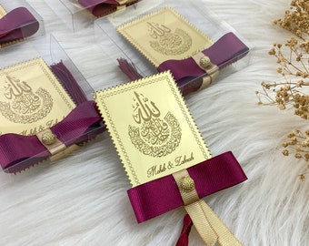 May include: Gold-colored card with Arabic calligraphy and the names "Mohib & Zahrah" printed on it. The card is decorated with a burgundy bow and a gold tassel.