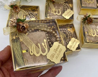 May include: Gold gift boxes with clear lids containing a small brown velvet book with a gold Arabic calligraphy design, a gold tassel, and a string of gold beads. Each box has a gold tag with the name "Ali's Ameen" and the date "10.22.2022".
