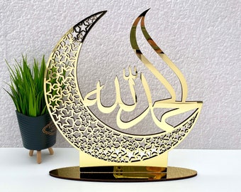 Op de afbeelding: Een goudkleurig decoratief stuk met een halve maanvorm met een sterrenpatroon en Arabische kalligrafie. Het stuk is gemonteerd op een cirkelvormige basis en heeft een reflecterend oppervlak. Een kleine plant in een pot is zichtbaar op de achtergrond.