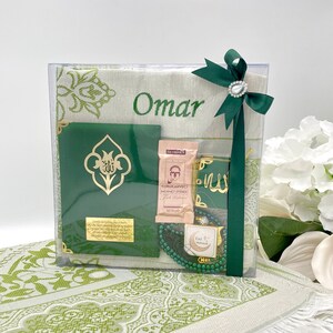 May include: A green and white prayer rug gift set with a personalized name "Omar" embroidered on the rug. The set includes a prayer rug, a green prayer book, a small green pouch, a string of green prayer beads, and a small box of tea.