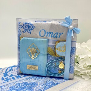 May include: A blue and white prayer rug set with a personalized name "Omar" embroidered on the rug. The set includes a prayer rug, a prayer mat, a small book, a prayer bead necklace, and a decorative tassel.
