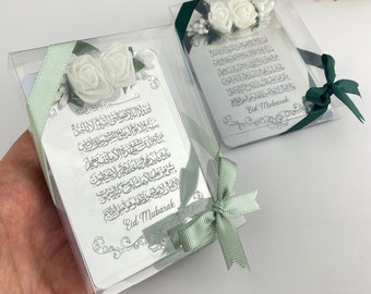 May include: Two silver gift boxes with green ribbon and white flowers. The boxes contain cards with Arabic calligraphy and the text "Eid Mubarak".