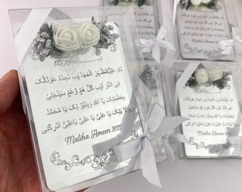 May include: Clear acrylic favors with silver text and floral accents. Each favor features a printed design with Arabic script, white roses, and a silver ribbon. The favors are packaged in clear boxes, perfect for special occasions.