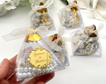 May include: Four clear pyramid-shaped boxes with gold accents, each containing a white pearl necklace and a gold mirror with the names "Yasmeen & Ibrahim" inscribed. The boxes are tied with white ribbon.