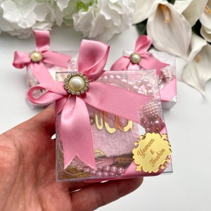 May include: Clear square gift boxes tied with pink satin ribbons and pearl-centered embellishments. Each box contains a pink prayer mat, a string of pink pearls, and a gold-colored tag with the names "Yasmeen & Ibrahim".