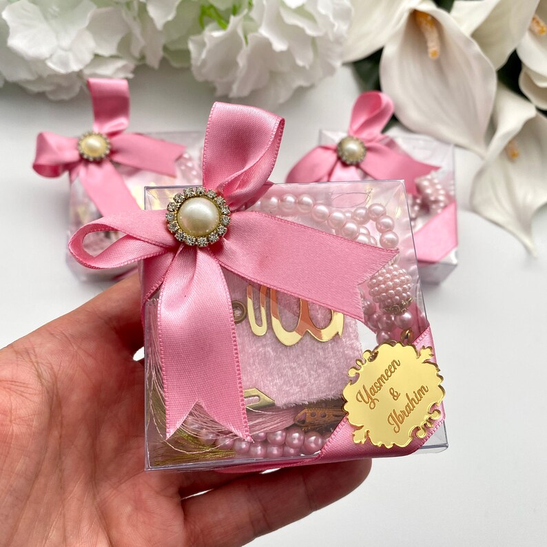 May include: Clear square gift boxes tied with pink satin ribbons and pearl-centered embellishments. Each box contains a pink prayer mat, a string of pink pearls, and a gold-colored tag with the names "Yasmeen & Ibrahim".