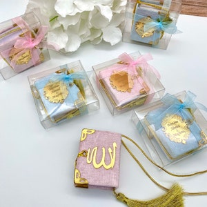 May include: A set of small, decorative, fabric-covered books with gold accents and tassels. The books are in pink and blue colours and have personalised names on them. The books are in clear plastic boxes with ribbons.