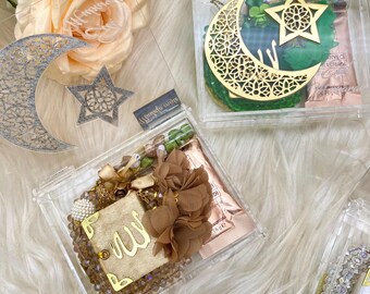 May include: Three clear acrylic boxes filled with decorative items. The boxes are filled with a variety of items, including a gold crescent moon and star, a brown flower, a gold charm with the word "Allah" written on it, and a small bottle of perfume. The boxes are labeled with the names "Ismahane" and "Alhamdulillah"