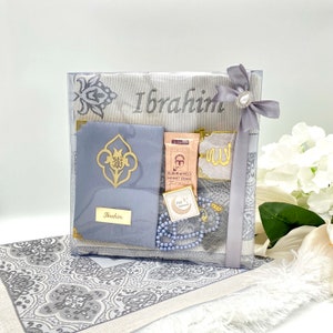 May include: A gray and white prayer rug gift set with a personalized name "Ibrahim" embroidered on the rug. The set includes a prayer rug, a small prayer mat, a prayer bead necklace, a small decorative box, and a small bag of Turkish coffee.