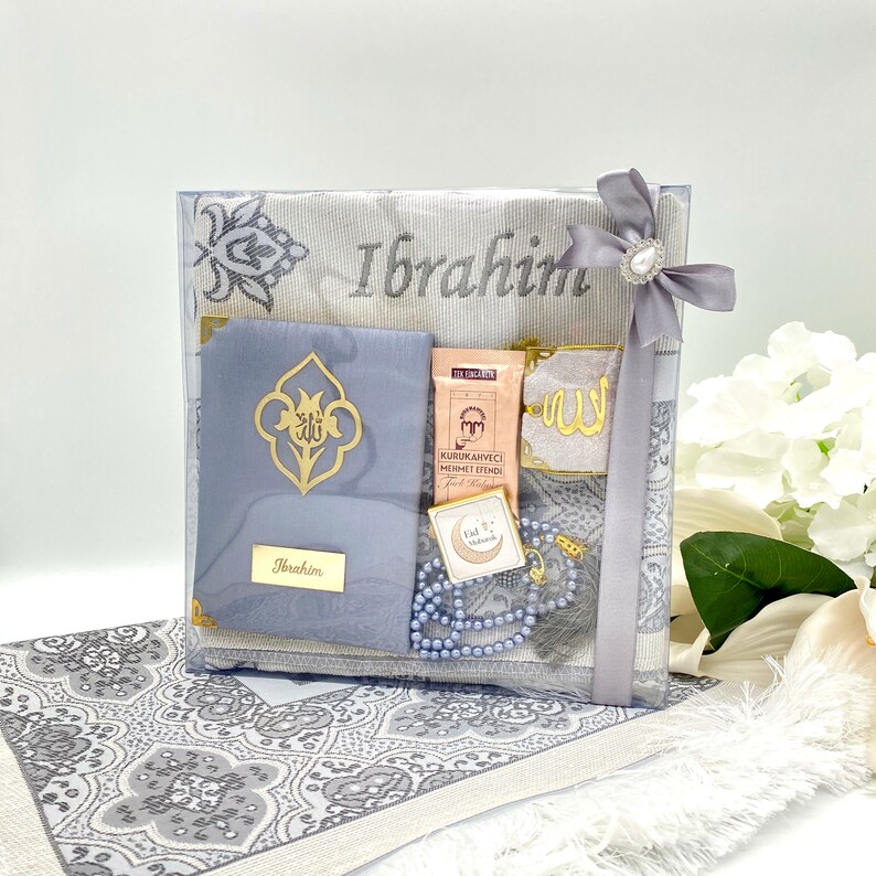 May include: A gray and white prayer rug gift set with a personalized name "Ibrahim" embroidered on the rug. The set includes a prayer rug, a small prayer mat, a prayer bead necklace, a small decorative box, and a small bag of Turkish coffee.