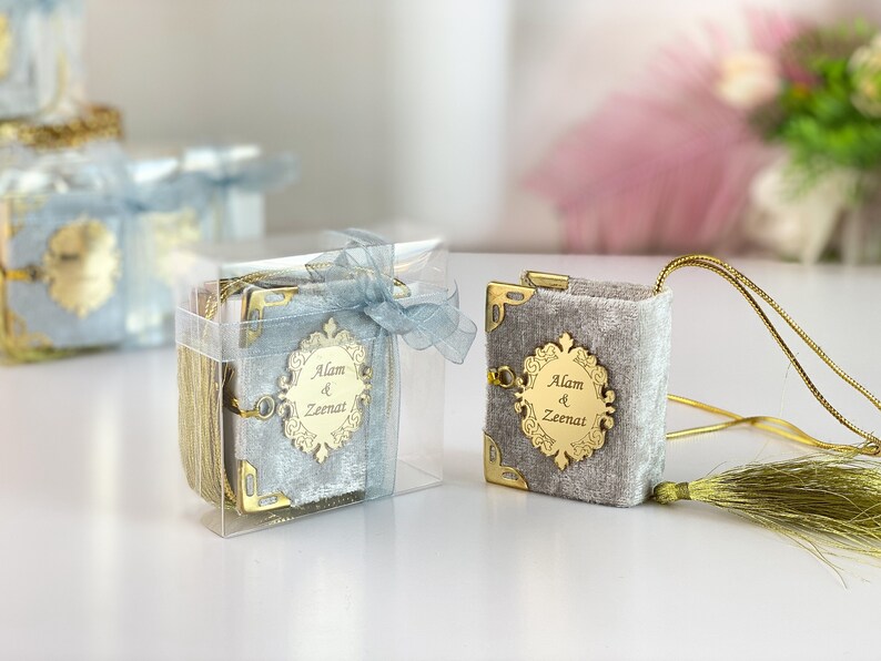 May include: Two small, decorative books with gold accents and a velvet cover. The books are personalised with the names "Alam & Zeenat". One book is in a clear plastic box with a blue ribbon, the other book has a gold chain and a green tassel.