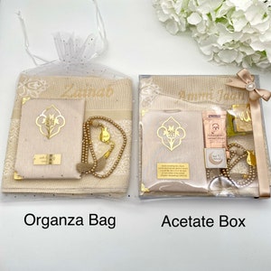 K&ouml;nnte beinhalten: Zwei Geschenksets zum Beten, eines in einer beigefarbenen Organza-Tasche und das andere in einer durchsichtigen Acetat-Box. Beide Sets enthalten ein Gebetsteppich, ein Gebetbuch, eine Gebetskette und ein Lesezeichen. Das Set in der Organza-Tasche ist mit dem Namen "Zainab" personalisiert und das Set in der Acetat-Box ist mit dem Namen "Ammi Jada" personalisiert.