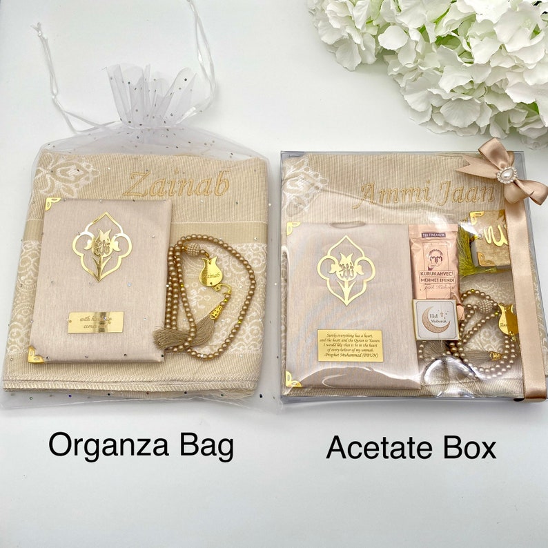 K&ouml;nnte beinhalten: Zwei Geschenksets zum Beten, eines in einer beigefarbenen Organza-Tasche und das andere in einer durchsichtigen Acetat-Box. Beide Sets enthalten ein Gebetsteppich, ein Gebetbuch, eine Gebetskette und ein Lesezeichen. Das Set in der Organza-Tasche ist mit dem Namen "Zainab" personalisiert und das Set in der Acetat-Box ist mit dem Namen "Ammi Jada" personalisiert.