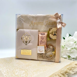 May include: A beige prayer rug gift set with a personalized embroidered prayer rug, a small prayer rug, a prayer bead necklace, a small box with a crescent moon and star design, a small box with a prayer rug design, and a small gold-colored plaque with the word "Allah" in Arabic script.