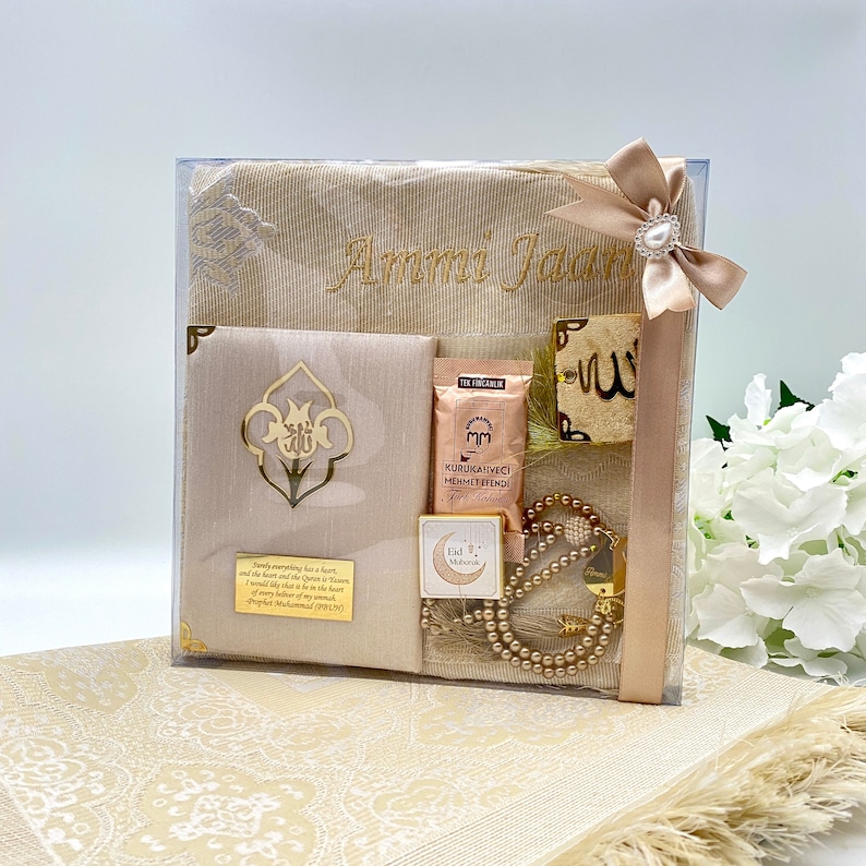 May include: A beige prayer rug gift set with a personalized embroidered prayer rug, a small prayer rug, a prayer bead necklace, a small box with a crescent moon and star design, a small box with a prayer rug design, and a small gold-colored plaque with the word "Allah" in Arabic script.