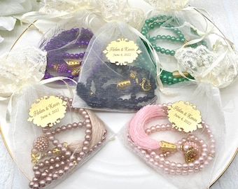 May include: Four white organza bags with gold trim, each containing a different colored prayer bead necklace. The necklaces are black, purple, pink, and green. Each bag has a gold tag with the text "Helen & Kevin June 4, 2022".