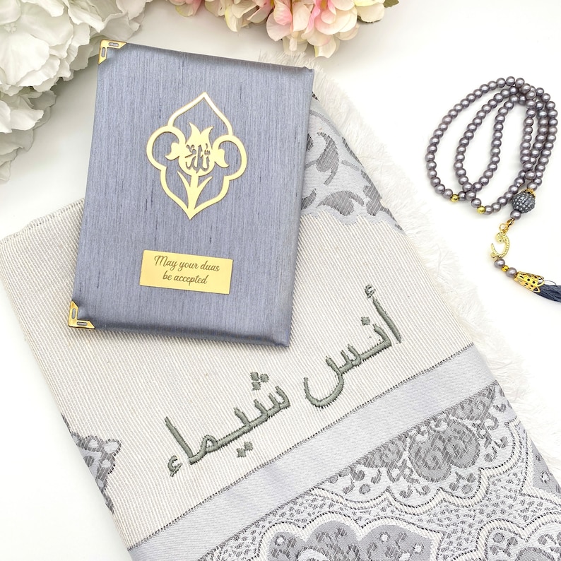 K&ouml;nnte beinhalten: Ein grauer Gebetsteppich mit einem silbernen und wei&szlig;en Blumenmuster. Der Teppich ist mit dem arabischen Satz "M&ouml;gen deine Duas angenommen werden" bestickt. Ein grauer Gebetsteppich mit einem goldenen Blumendesign und dem Satz "M&ouml;gen deine Duas angenommen werden" ist enthalten. Eine graue Gebetskette mit einem goldenen Halbmond-Anh&auml;nger ist ebenfalls enthalten.