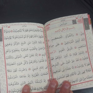 Pu&ograve; includere: Un libro aperto con testo arabo in rosso e nero. Il libro &egrave; aperto a una pagina con il titolo "Sura Yasin" e il testo "Bismillah ir-Rahman ir-Rahim".
