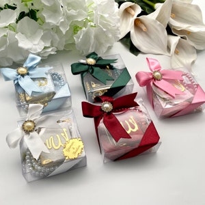 May include: Several clear boxes with gifts, each tied with a satin ribbon and a pearl-centered embellishment. The boxes contain prayer beads and small books. Ribbons are in shades of blue, green, pink, white, and burgundy. White flowers are in the background.