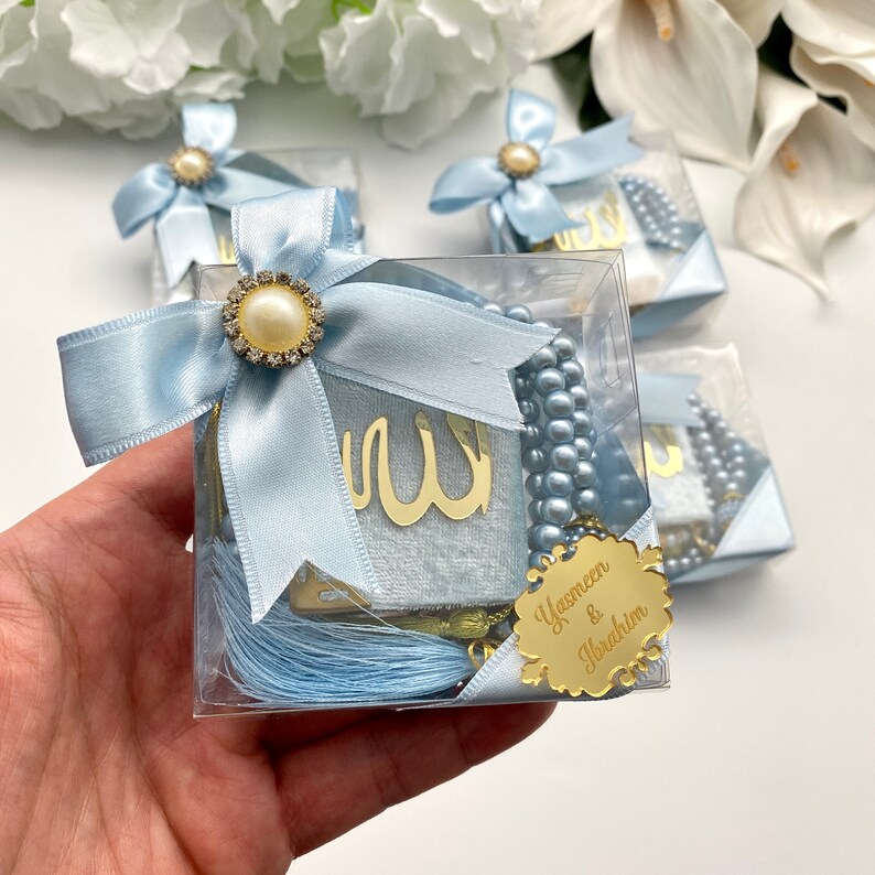 May include: Clear square boxes tied with light blue satin bows, each containing a small book with gold Arabic script, a string of beads, and a tassel. The boxes are decorated with a gold name tag and a pearl and rhinestone embellishment.