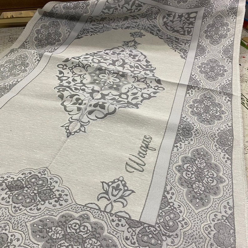 May include: A white prayer rug with a gray floral pattern and the word "Wajigas" in the center.