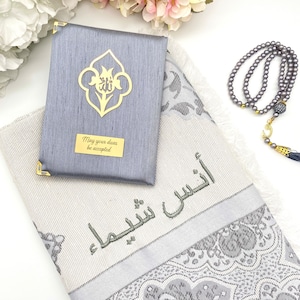 Puede incluir: Una alfombra de oraci&oacute;n gris con un patr&oacute;n floral plateado y blanco. La alfombra tiene bordada la frase &aacute;rabe "Que tus du'as sean aceptadas". Se incluye una alfombra de oraci&oacute;n gris con un dise&ntilde;o floral dorado y la frase "Que tus du'as sean aceptadas". Tambi&eacute;n se incluye un collar de cuentas de oraci&oacute;n gris con un dije de media luna dorada.