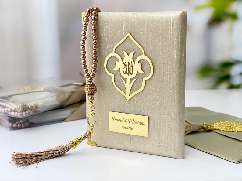 Pu&ograve; includere: Un libro di preghiere beige con un motivo floreale dorato e l'iscrizione "Navid & Mariam 29/03/2022". Un fiocco marrone e una collana di preghiera con perline marroni sono attaccati al libro.