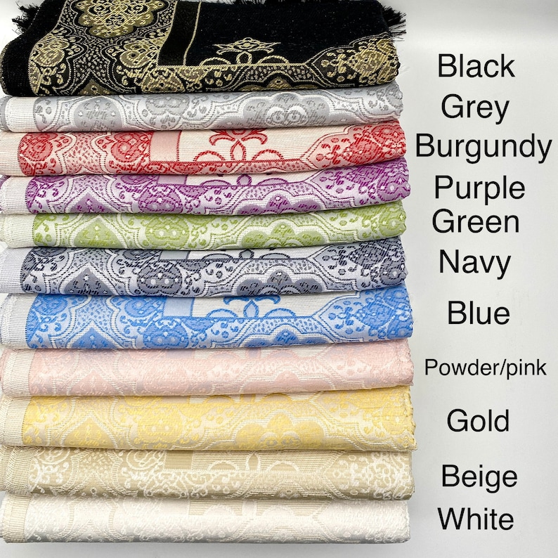 May include: A stack of ten prayer rugs in various colors, including black, grey, burgundy, purple, green, navy, blue, powder pink, gold, beige, and white. The rugs are made of a soft, textured fabric and feature intricate patterns.