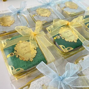May include: A collection of small, green velvet books with gold accents and a gold inscription that reads "Alam & Zeenat". The books are packaged in clear plastic boxes with gold ribbon and blue ribbon.