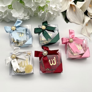 May include: Clear square boxes tied with satin ribbons in various colors, each adorned with a pearl-centered embellishment. Inside, there are prayer beads, small books with gold lettering, and decorative tassels. The ribbons are in shades of blue, green, white, burgundy, and pink.