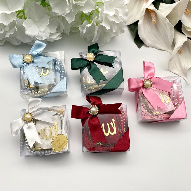 May include: Clear square boxes tied with satin ribbons in various colors, each adorned with a pearl-centered embellishment. Inside, there are prayer beads, small books with gold lettering, and decorative tassels. The ribbons are in shades of blue, green, white, burgundy, and pink.