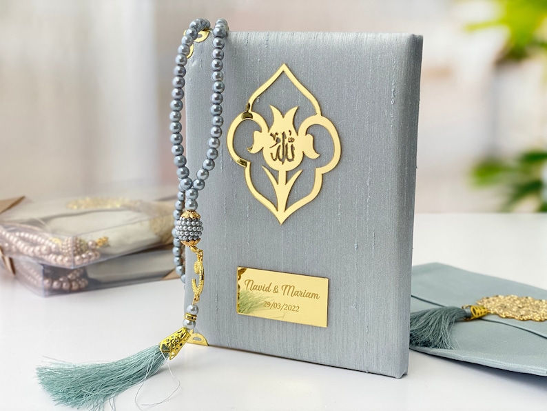 Pu&ograve; includere: Un libro di preghiere in velluto grigio con un motivo floreale dorato e la parola "Allah" in scrittura araba. Il libro ha un'iscrizione personalizzata che recita "David & Mariam 29/03/2022". Viene fornito con un fiocco e grani di preghiera abbinati di colore grigio.