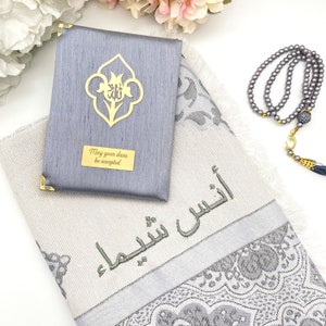 May include: A gray prayer rug with a silver and white floral pattern. The rug has the Arabic phrase "May your duas be accepted" embroidered on it. A gray prayer mat with a gold floral design and the phrase "May your duas be accepted" is included. A gray prayer bead necklace with a gold crescent moon charm is also included.
