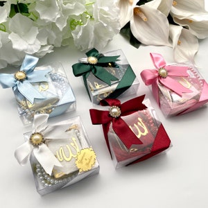 May include: Five clear square gift boxes with satin bows in blue, green, pink, white, and burgundy. Each box is adorned with a pearl and rhinestone embellishment. The boxes contain prayer beads and a gold-colored decorative element with Arabic script.