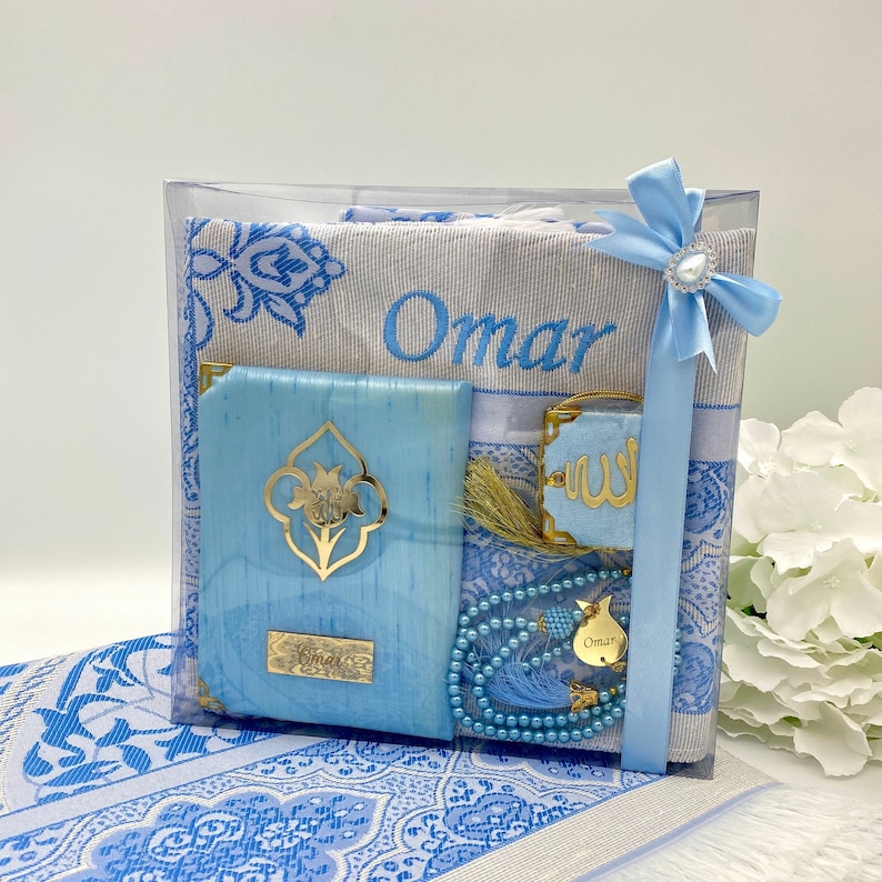 May include: A blue and white prayer rug set with a personalized name "Omar" embroidered on the rug. The set includes a prayer rug, a prayer mat, a small book, a prayer bead necklace, and a decorative tassel.