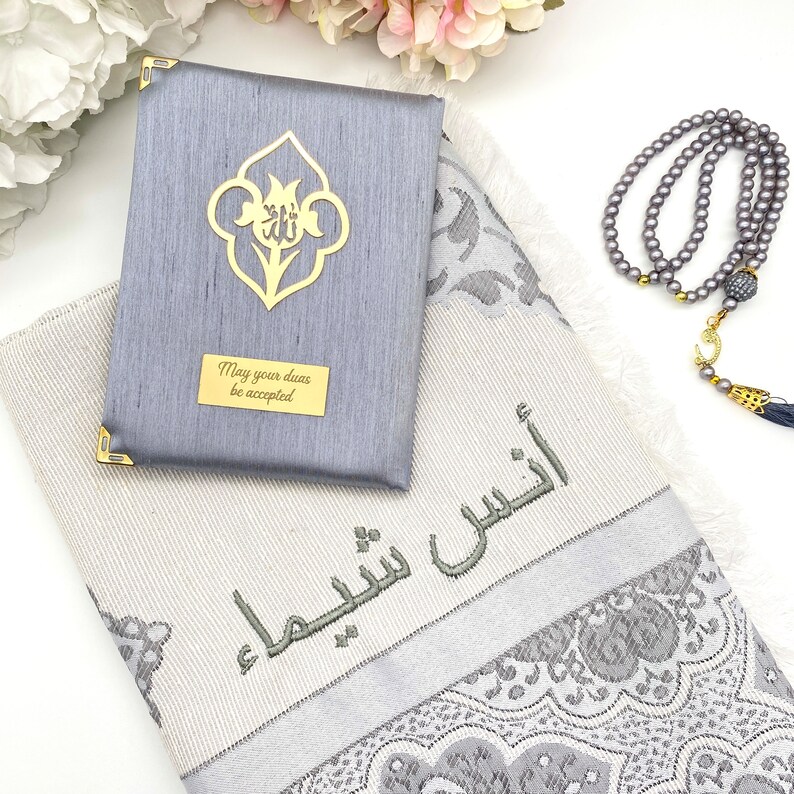 May include: A gray prayer rug with a silver and white floral pattern. The rug has the Arabic phrase "May your duas be accepted" embroidered on it. A gray prayer mat with a gold floral design and the phrase "May your duas be accepted" is included. A gray prayer bead necklace with a gold crescent moon charm is also included.